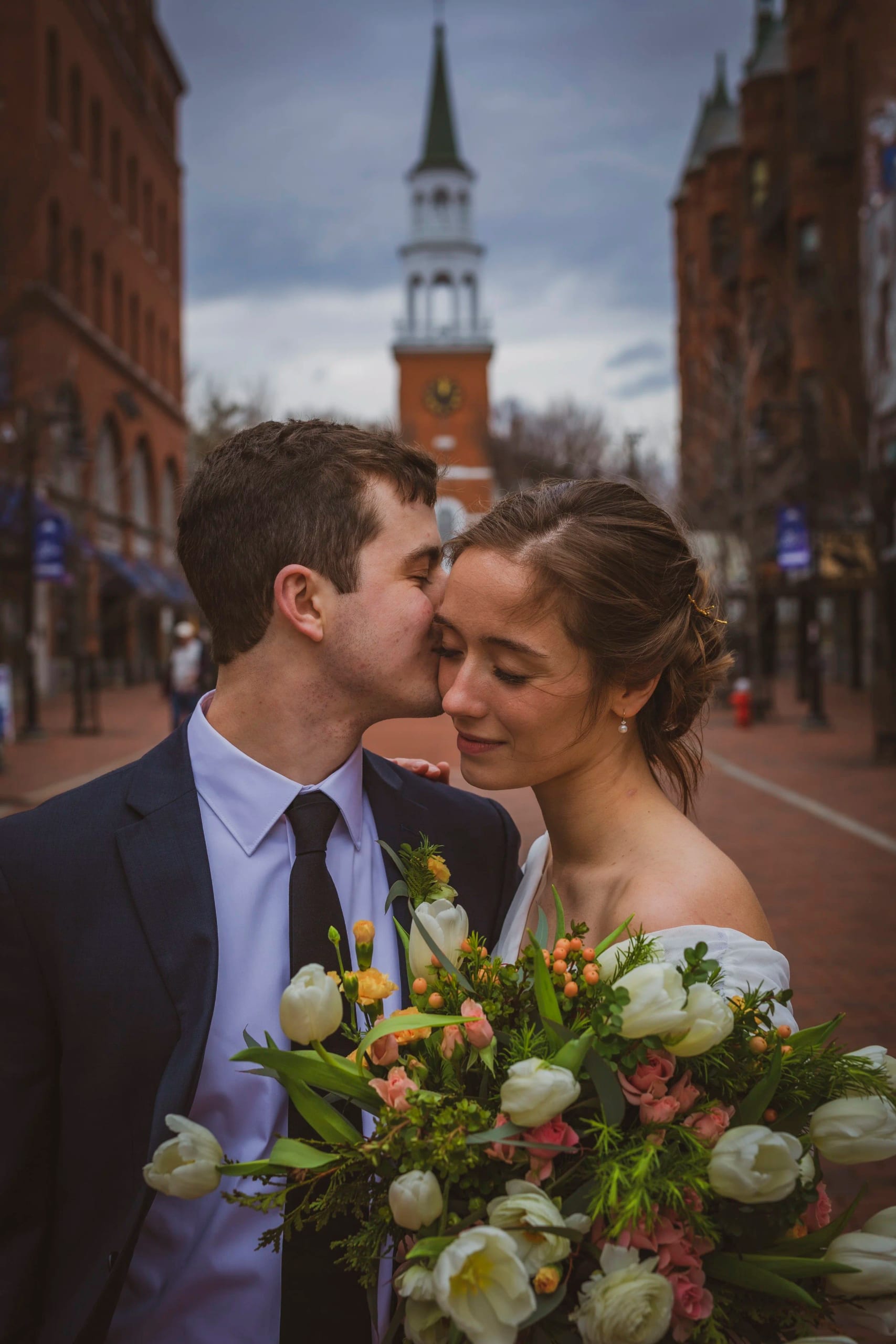 A couple shares a kiss on a brick street with a white church steeple behind them, the bride holding a bouquet of tulips