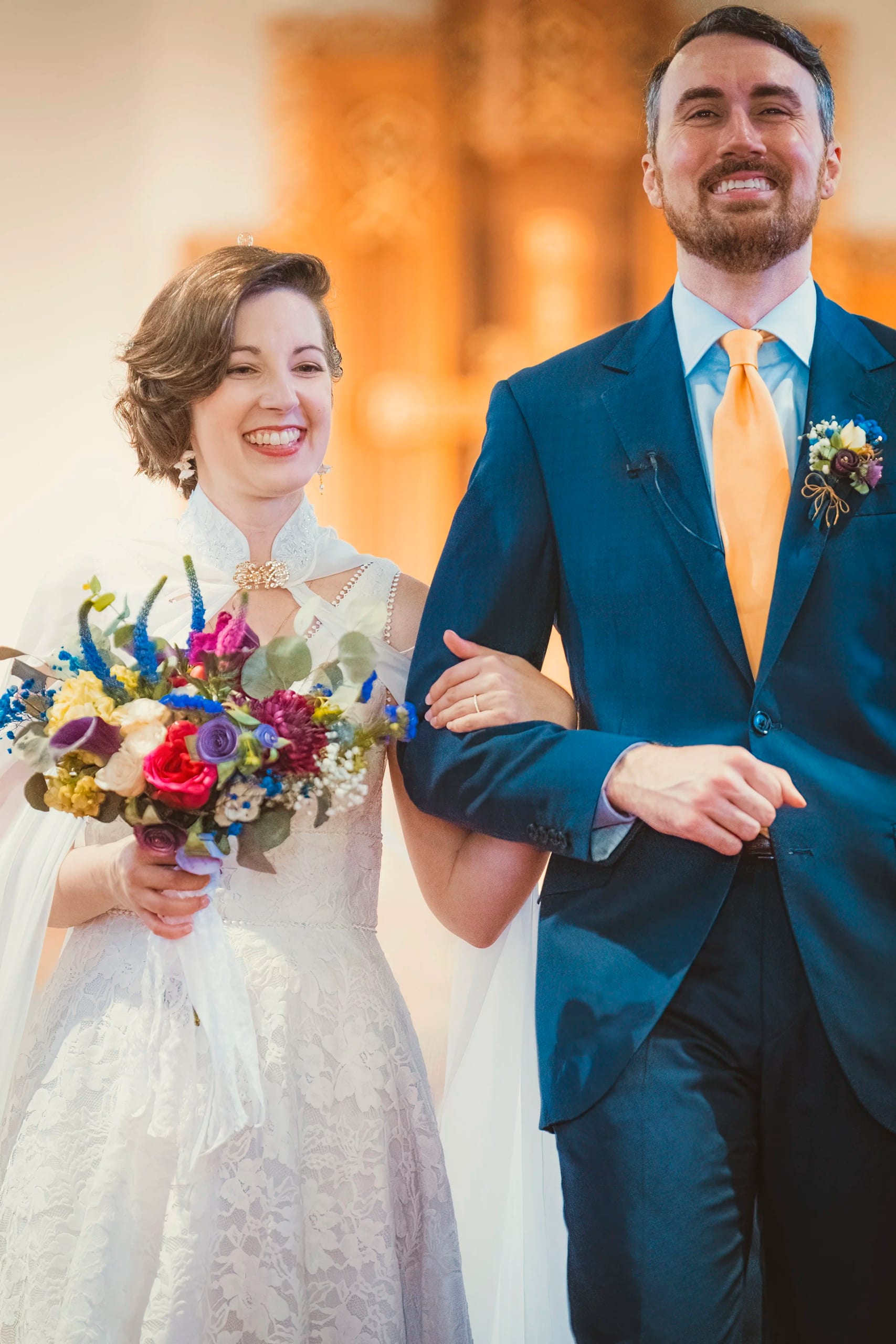 A smiling bride and groom walk arm in arm after their ceremony, her bouquet of colorful flowers in hand