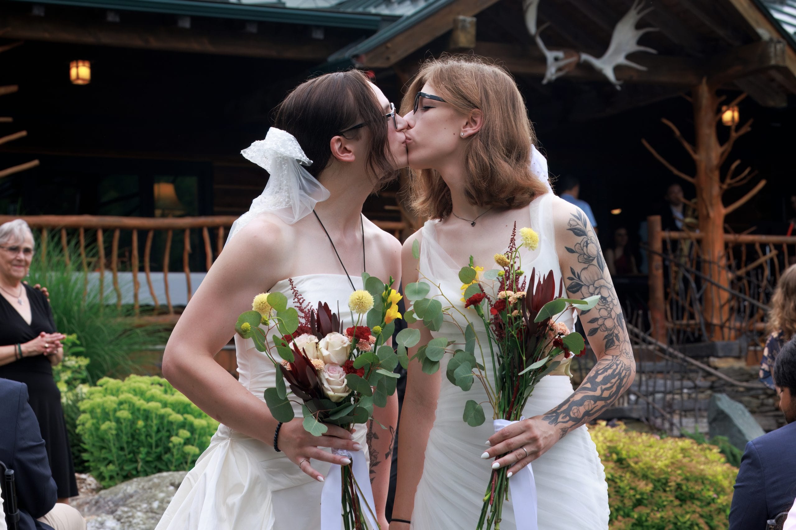 Two brides in white dresses share a kiss while holding bouquets at an outdoor ceremony