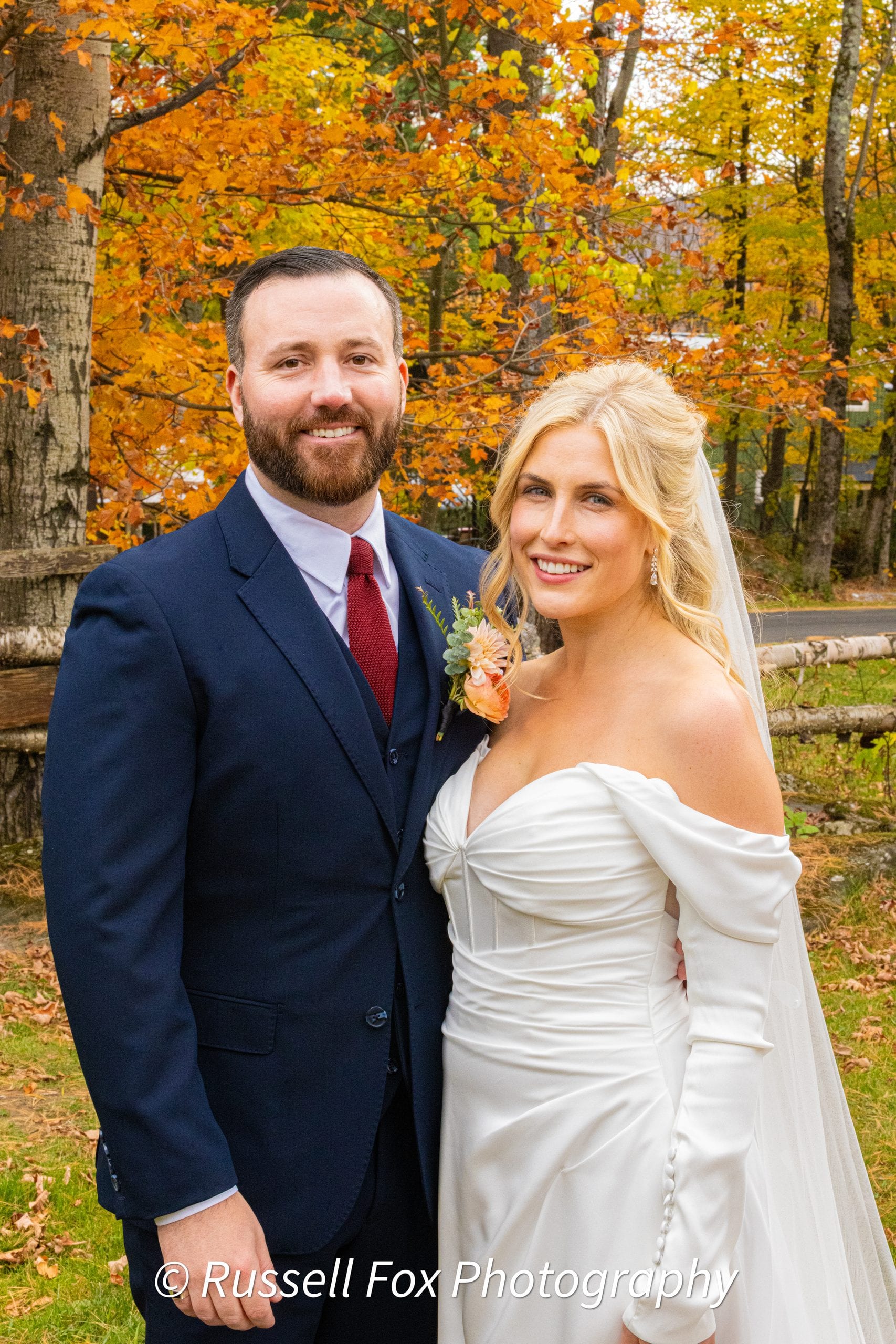 A smiling bride and groom pose together in front of autumn foliage