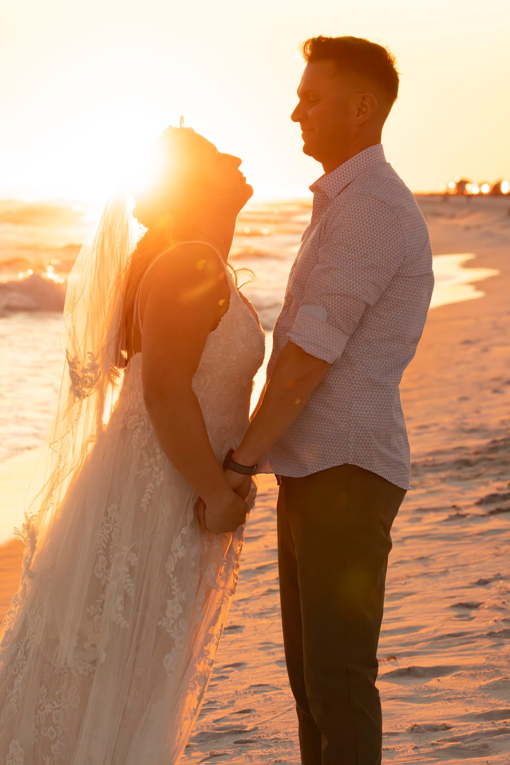 A bride and groom stand hand in hand on the beach at sunset