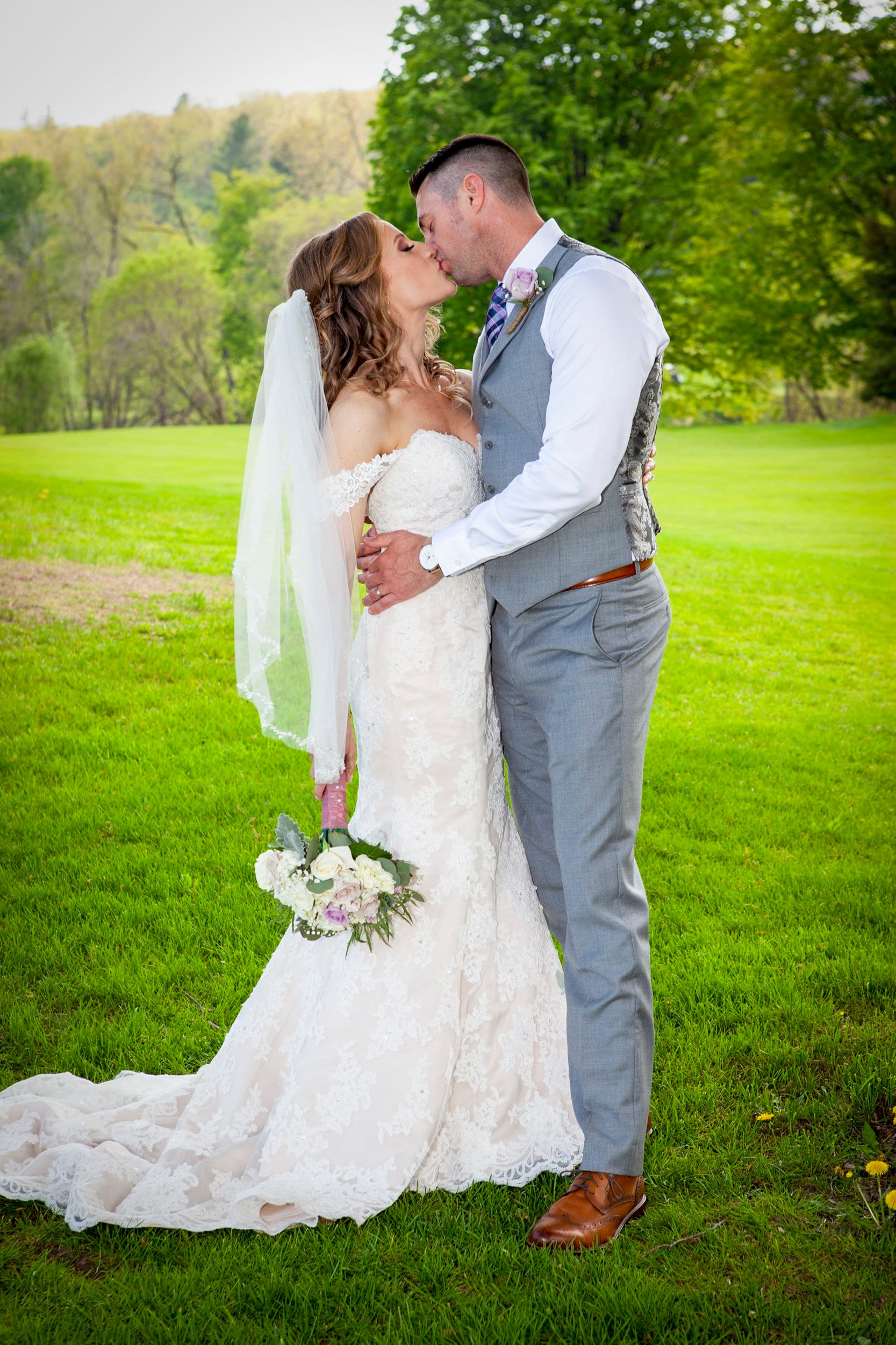A bride and groom kiss on a green lawn, her veil blowing behind her