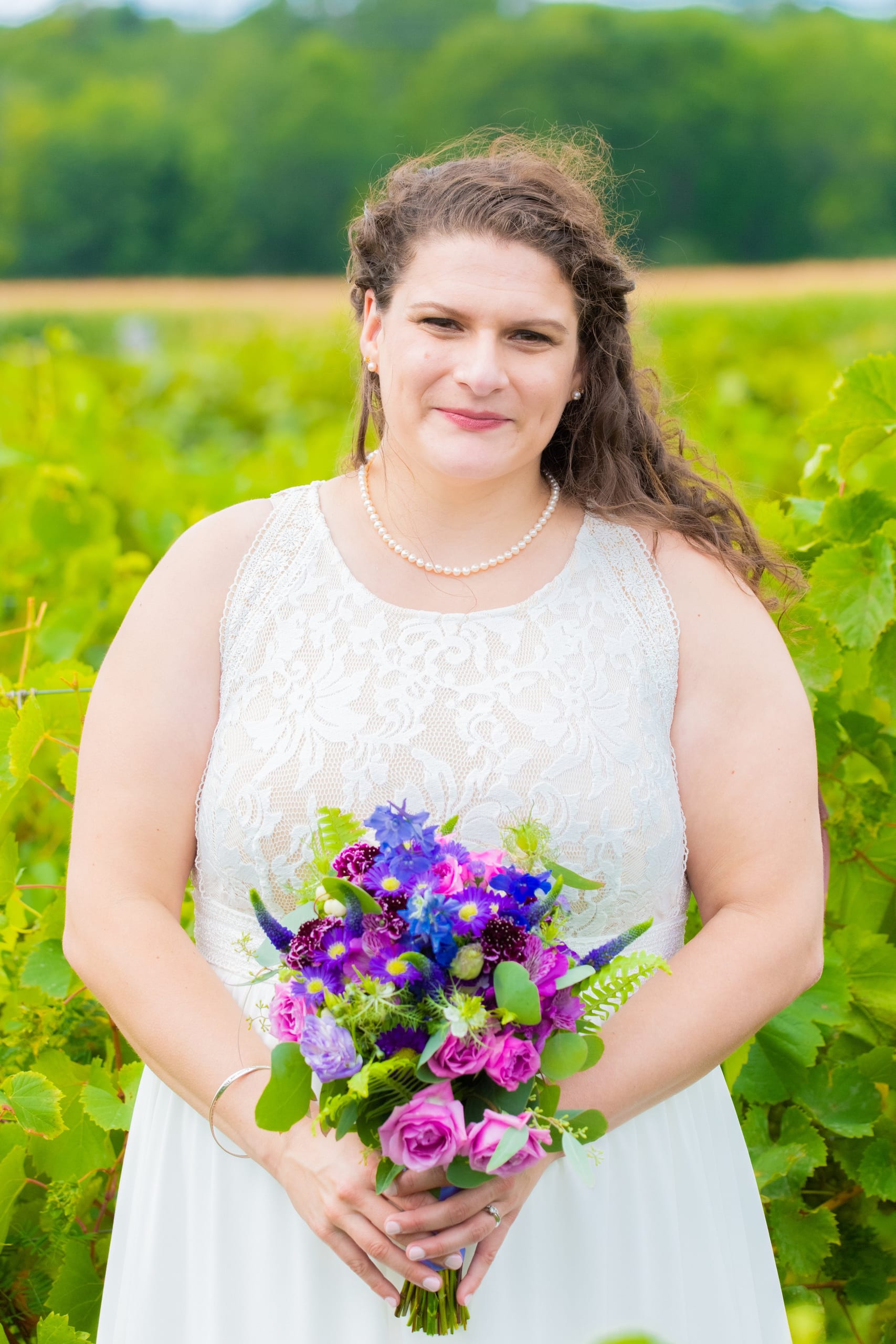 A bride holding a purple and pink bouquet in front of the vineyard rows