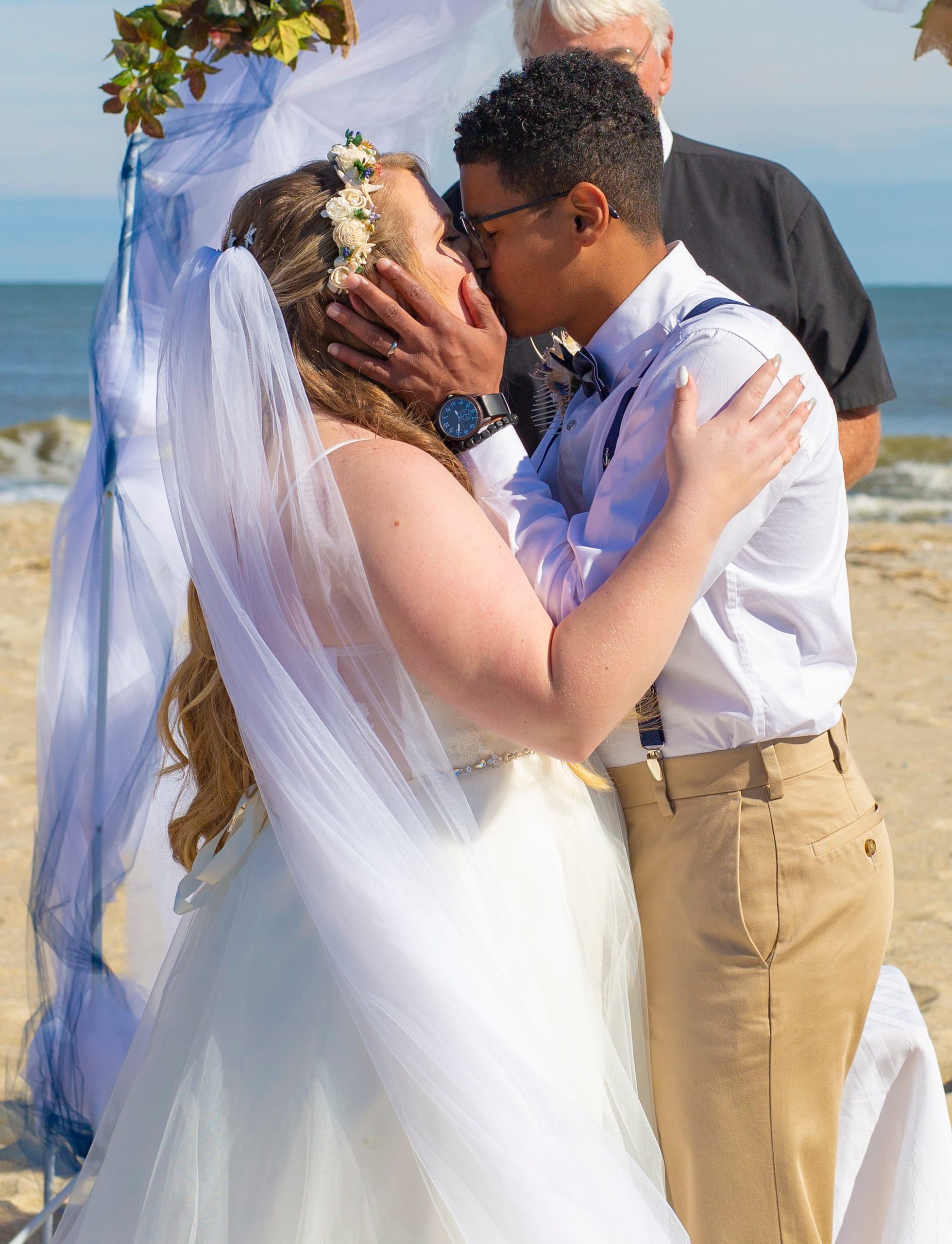A bride and groom kiss under a beach ceremony arch with the ocean behind them
