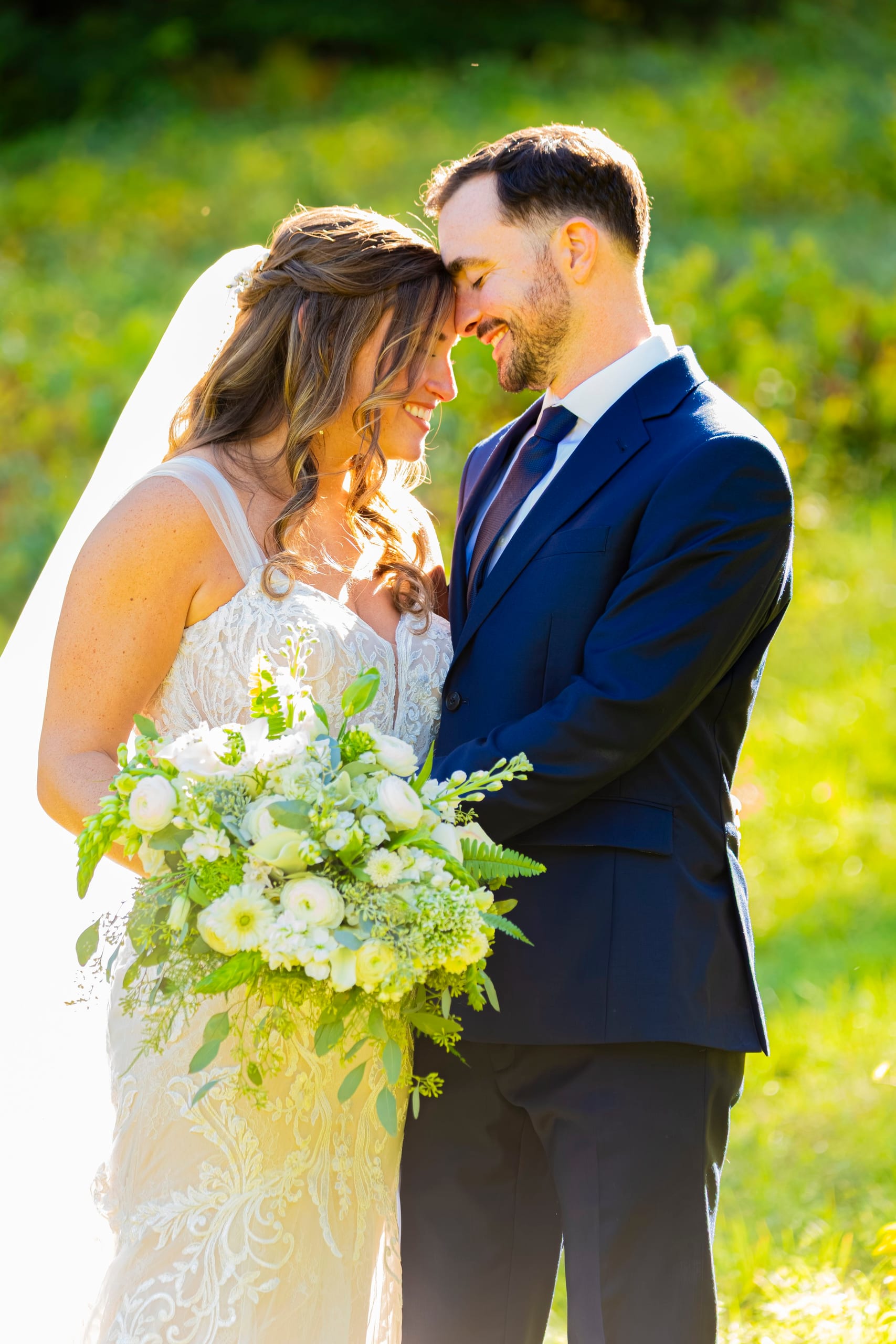 A bride and groom touch foreheads in golden light, she holds a white bouquet